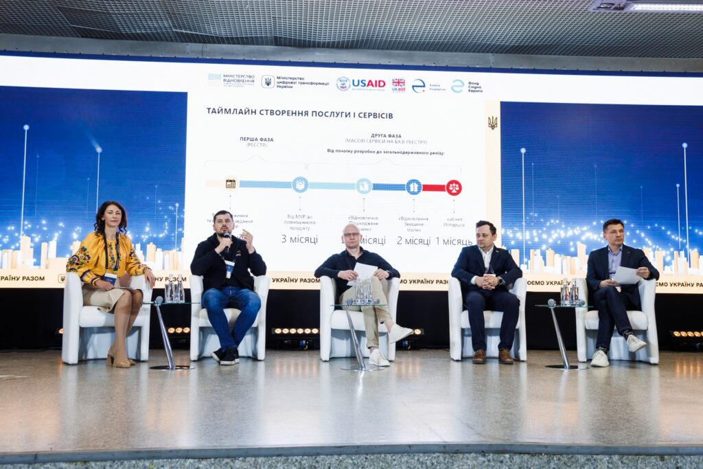 Five people sit on stage in front of a large screen wall. The screen displays information about eRecovery and the logos of USAID, UK Aid, Ukraine's MCTID, MDT, Eurasia Foundation, and the East Europe Foundation.