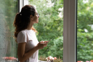 A young white woman with curly brown hair and glasses stands looking out a window, with her profile toward the viewer. She gazes at the greenery outside the window while holding a smartphone in her hands.