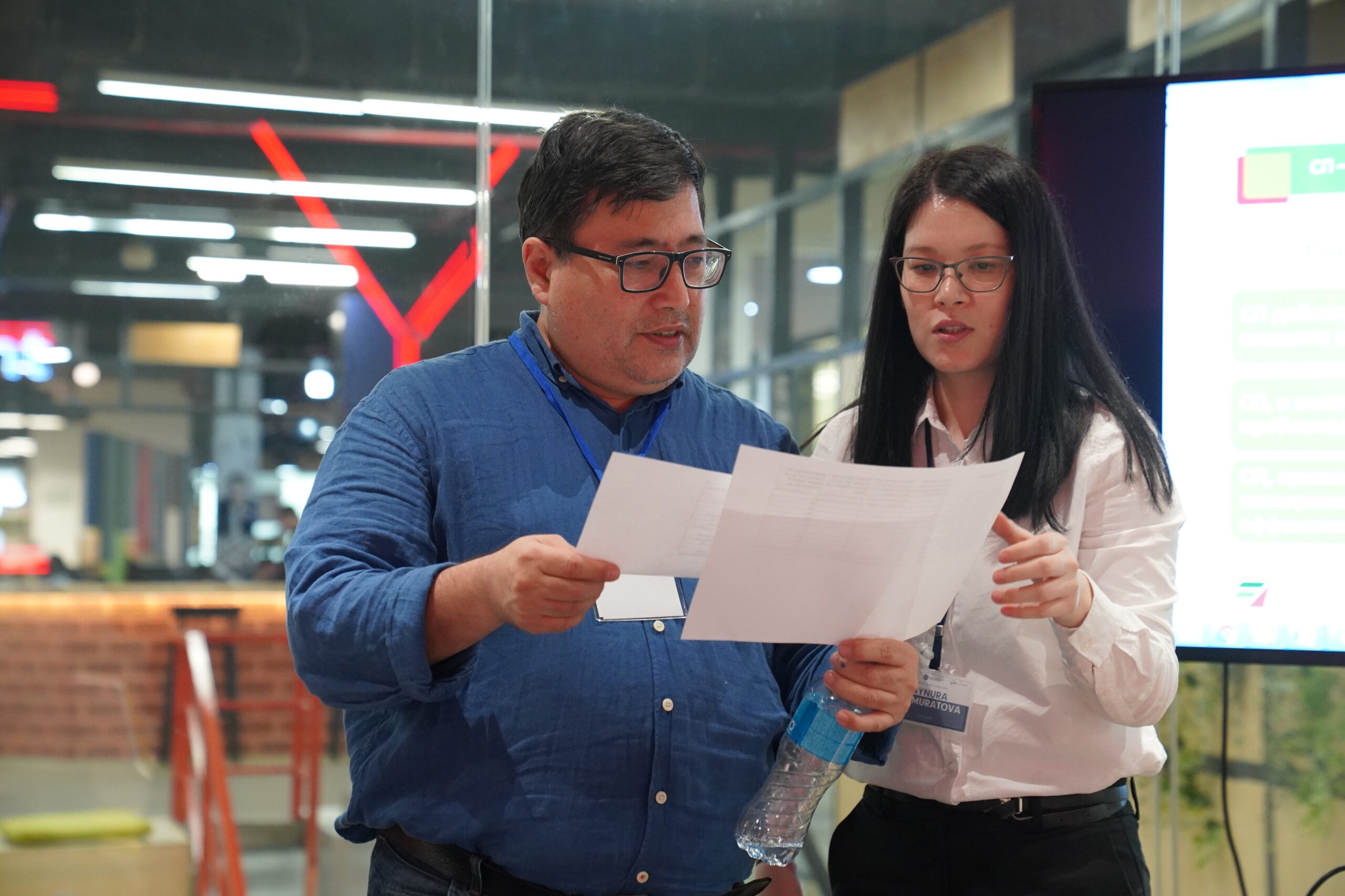 Two people review documents. To the left, a man in a blue shirt holds two pieces of paper. To his right, a woman in a white shirt reaches for a paper and reads alongside, in apparent discussion.